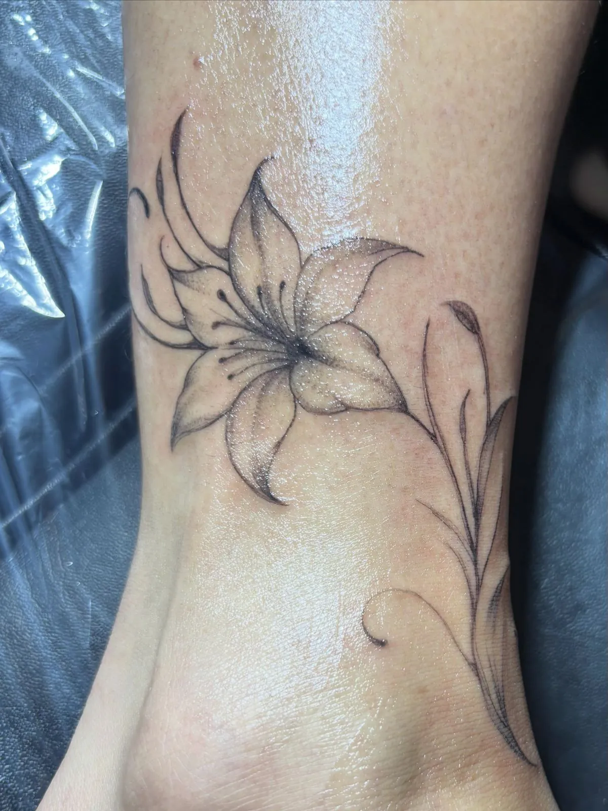 Fine line lily tattoo with long flowing stems on an ankle