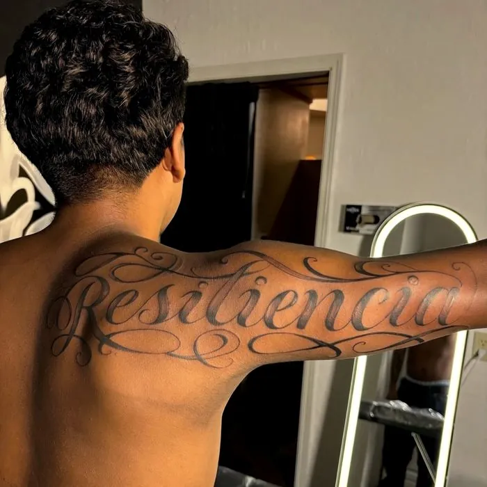 Large script lettering tattoo reading Resilencia across the shoulders. View on Instagram.