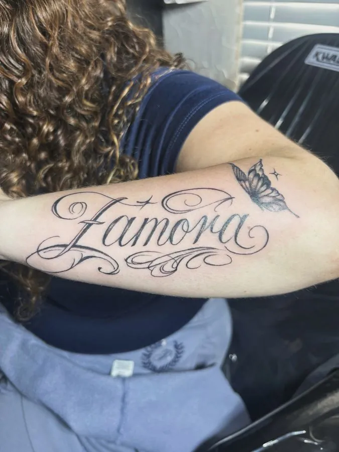 Script name tattoo with a small butterfly on the forearm